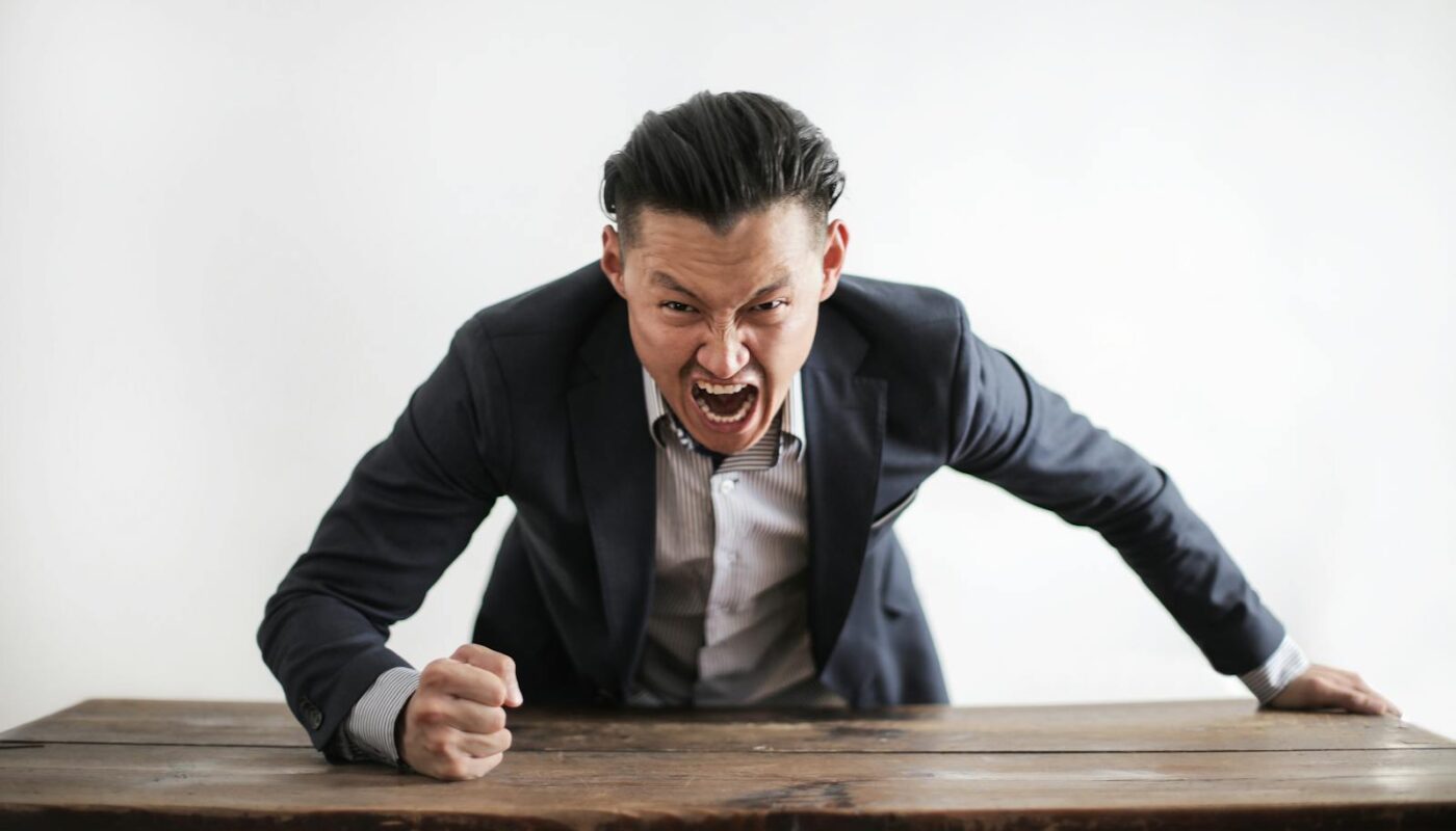 Homme d'affaires expressif en colère en costume formel regardant la caméra et criant de folie tout en frappant le bureau avec le poing