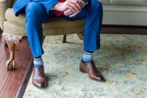 Gros plan sur les jambes d'un homme en costume bleu et chaussures en cuir marron, portant des chaussettes fantaisie homme à rayures bleues et blanches, assis dans un fauteuil élégant.