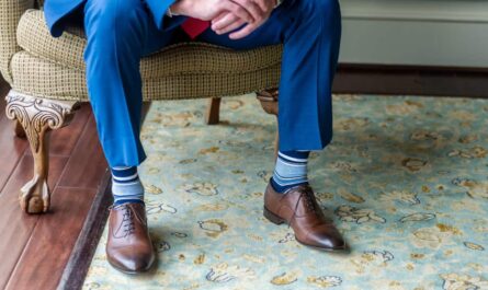 Gros plan sur les jambes d'un homme en costume bleu et chaussures en cuir marron, portant des chaussettes fantaisie homme à rayures bleues et blanches, assis dans un fauteuil élégant.